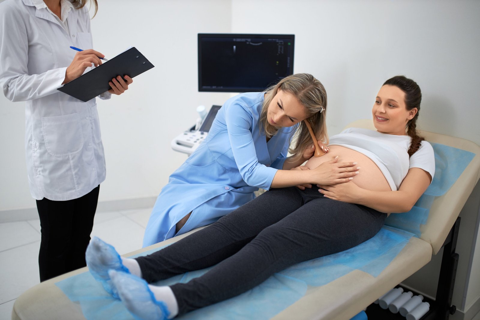 Professional female gynecologist checking pregnant woman's tummy with special medical equipment. Assistant in white lab coat standing near and writing in clipboard.